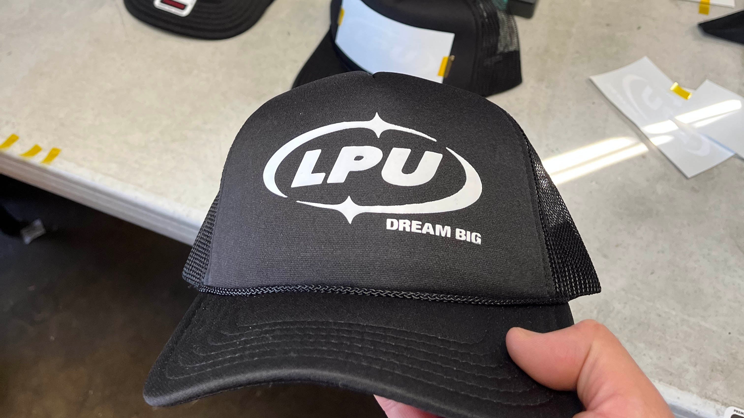 Black cap with 'LPU' logo held by a hand, with a teal cap size chart in the background.
